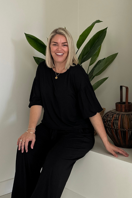 Woman wearing the Lexi Bamboo Top in black styled with Alexa Pants in black, smiling and seated indoors.