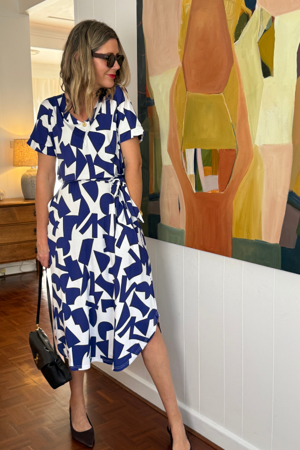Model showcasing Natali The Label's Bella Dress in Navy Abstract bamboo fabric—featuring striking geometric blue and white patterns, styled with black sunglasses and handbag, set against a modern interior with abstract artwork and warm wood accents.