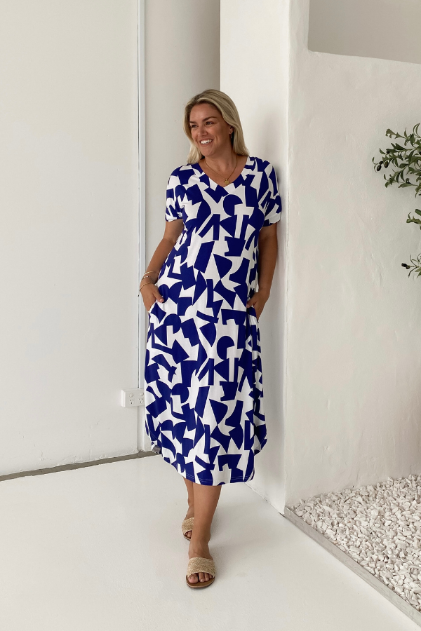 Model wearing the Bella Bamboo Tee Dress in navy abstract, hands casually in pockets, leaning against a white wall—showcasing the dress’s relaxed fit and laid-back elegance.