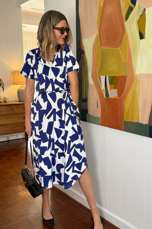 Woman wearing Natali The Label's Bella Dress in Navy Abstract print—crafted from sustainable bamboo fabric with bold blue and white geometric patterns, belted waist, and elegant silhouette, styled with black accessories in a modern indoor setting.